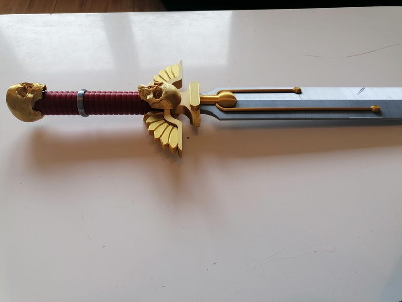 Op de afbeelding: Een gedetailleerd zwaard met een goudkleurige schedel op de pommel en de pareerstang. De handgreep is omwikkeld met rood materiaal. Het lemmet is zilverkleurig met gouden accenten. Fantasy-artikel.