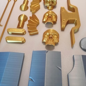 Op de afbeelding: Een verzameling goudkleurige 3D-geprinte objecten, waaronder schedels, decoratieve elementen en componenten voor een grotere structuur. Verschillende blauwgrijze panelen suggereren een project of model in aanbouw.