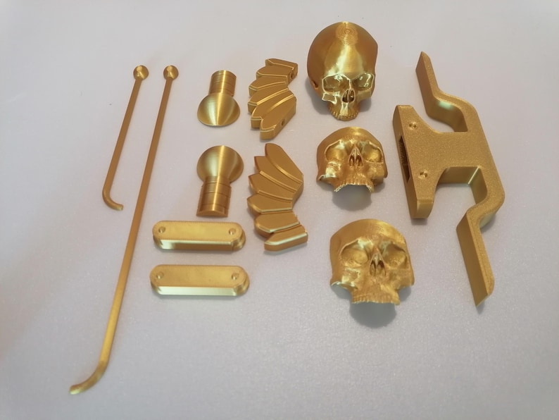 Op de afbeelding: Een verzameling goudkleurige 3D-geprinte objecten, waaronder drie schedelbeeldjes, vleugelvormige stukken en diverse geometrische vormen. Twee lange, dunne staven met afgeronde uiteinden en een gebogen haak zijn ook aanwezig.
