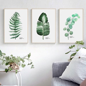 以下が含まれることがあります： 緑の葉を描いた水彩画が3枚、額装されています。絵は白い壁に飾られており、シダ、バナナの葉、ユーカリの枝が描かれています。それぞれの絵には「GROW」という言葉が書かれています。