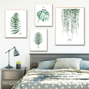以下が含まれることがあります： 白い背景に緑の葉とツルを描いた水彩画が4枚、額縁に入っています。絵は白い壁に掛けられており、その下には灰色のヘッドボードと緑と白の模様の掛け布団が敷かれたベッドがあります。