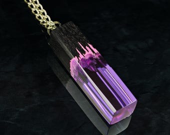 May include: A pendant necklace featuring a rectangular prism shape made of dark wood and translucent purple resin. The wood has a jagged edge where it meets the clear, light-catching purple resin. The pendant hangs from a thin gold-toned metal chain.