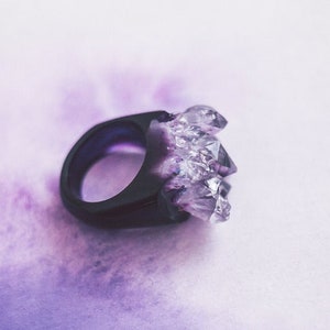 May include: A black ring featuring a cluster of clear, faceted crystals. The crystals sparkle, creating a striking effect. The ring is set against a soft, purple-toned background.