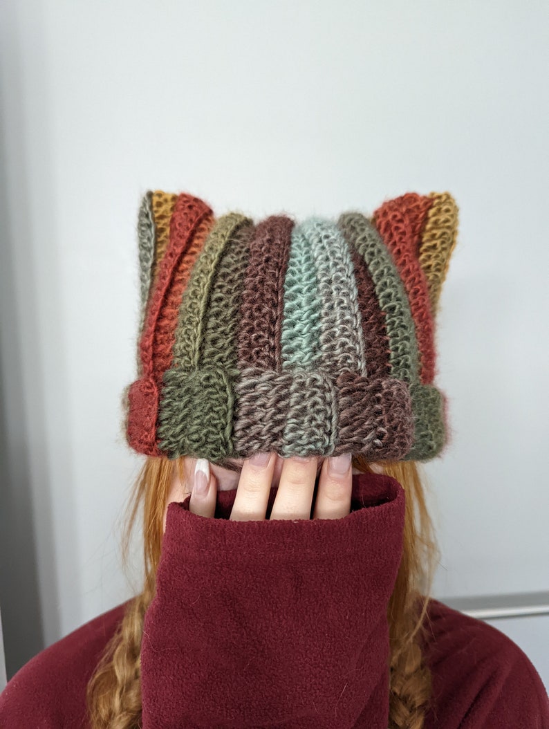 Puede incluir: Un gorro de crochet colorido con orejas de gato. El gorro est&aacute; hecho con una variedad de tonos de marr&oacute;n, verde y naranja. El gorro lo lleva una persona con el pelo largo y rojo.