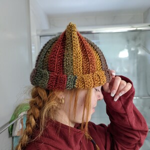 Puede incluir: Un gorro de punto de ganchillo multicolor con una parte superior puntiaguda. El gorro est&aacute; hecho con rayas de hilo marr&oacute;n, naranja, amarillo y verde.