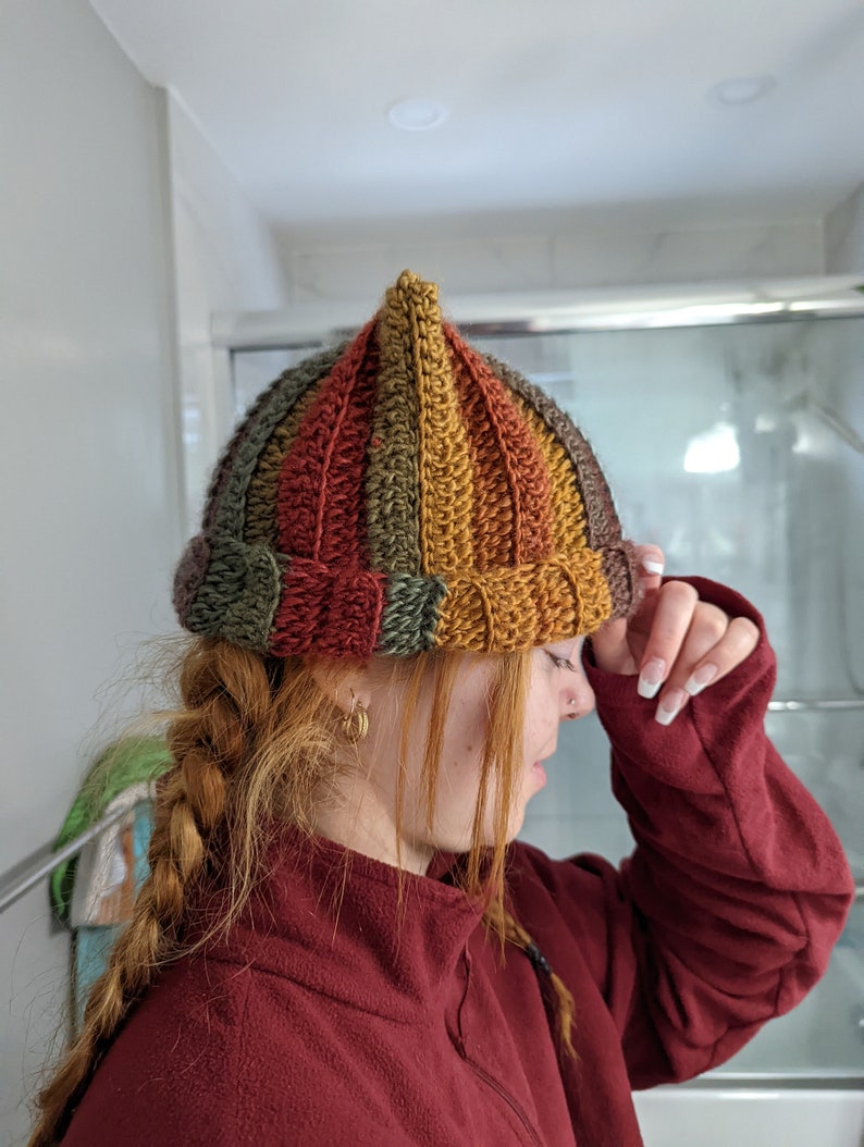 Puede incluir: Un gorro de punto de ganchillo multicolor con una parte superior puntiaguda. El gorro est&aacute; hecho con rayas de hilo marr&oacute;n, naranja, amarillo y verde.