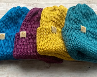 May include: Four knitted beanies in different colors: teal, burgundy, mustard yellow, and a darker teal. Each beanie has a small brown leather patch with a mountain logo.