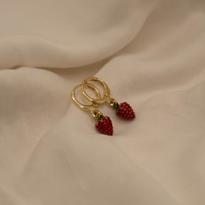 Könnte beinhalten: Ein Paar goldfarbene Creolen mit roten Erdbeer-Anhängern. Die Erdbeeren haben grüne Spitzen und sind mit winzigen roten Punkten versehen. Die Ohrringe werden auf einem cremefarbenen Stoff präsentiert.