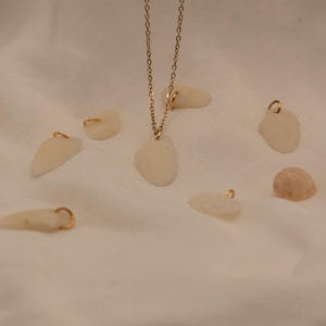 Könnte beinhalten: Eine goldene Halskette mit einem Anhänger aus einem glatten, cremefarbenen Stein. Mehrere ähnliche Steine mit Goldschleifen sind um die Halskette herum verstreut. Eine kleine, hellbraune Muschel ist ebenfalls vorhanden.