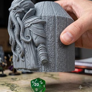 Puede incluir: Una torre de dados gris impresa en 3D que presenta una figura esculpida similar a un cefalópodo con tentáculos en la parte frontal y un cuerpo cilíndrico texturizado. Debajo de la torre hay un dado verde translúcido de veinte caras.