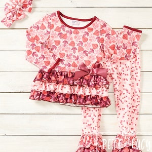 May include: A pink and burgundy two-piece outfit for a child. The top has a leaf print and a ruffled bottom. The pants have a polka dot print and a ruffled hem. The outfit comes with a matching headband.