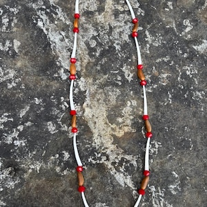 Puede incluir: Collar hecho a mano con un cordón blanco adornado con cuentas rojas y cuentas de madera alternas. El collar se exhibe sobre una superficie de piedra gris texturizada, mostrando sus elementos naturales.