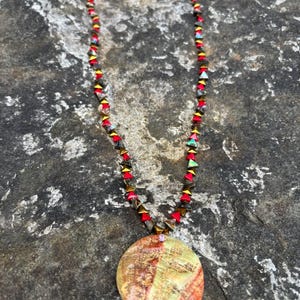 Pode incluir: Um colar artesanal com um pingente grande e redondo com um padrão marmorizado em tons de laranja, amarelo e vermelho. O colar tem uma corrente de contas com detalhes em vermelho, dourado e turquesa. O colar é exibido em uma superfície de pedra cinza.