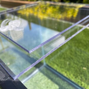 May include: Clear, rectangular glass panels, possibly for a terrarium or display case, are stacked. The edges of the glass have a slight purple tint. The background shows green grass and foliage, suggesting an outdoor setting. The glass reflects the surroundings.