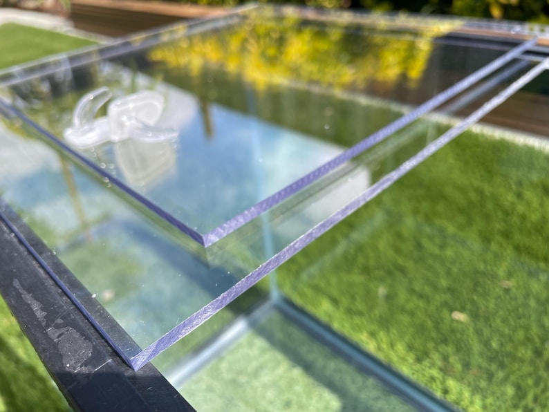 May include: Clear, rectangular glass panels, possibly for a terrarium or display case, are stacked. The edges of the glass have a slight purple tint. The background shows green grass and foliage, suggesting an outdoor setting. The glass reflects the surroundings.