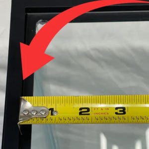 May include: A black picture frame with a clear glass pane. A yellow tape measure shows the frame's width is approximately 3 inches. The frame is on a white surface.