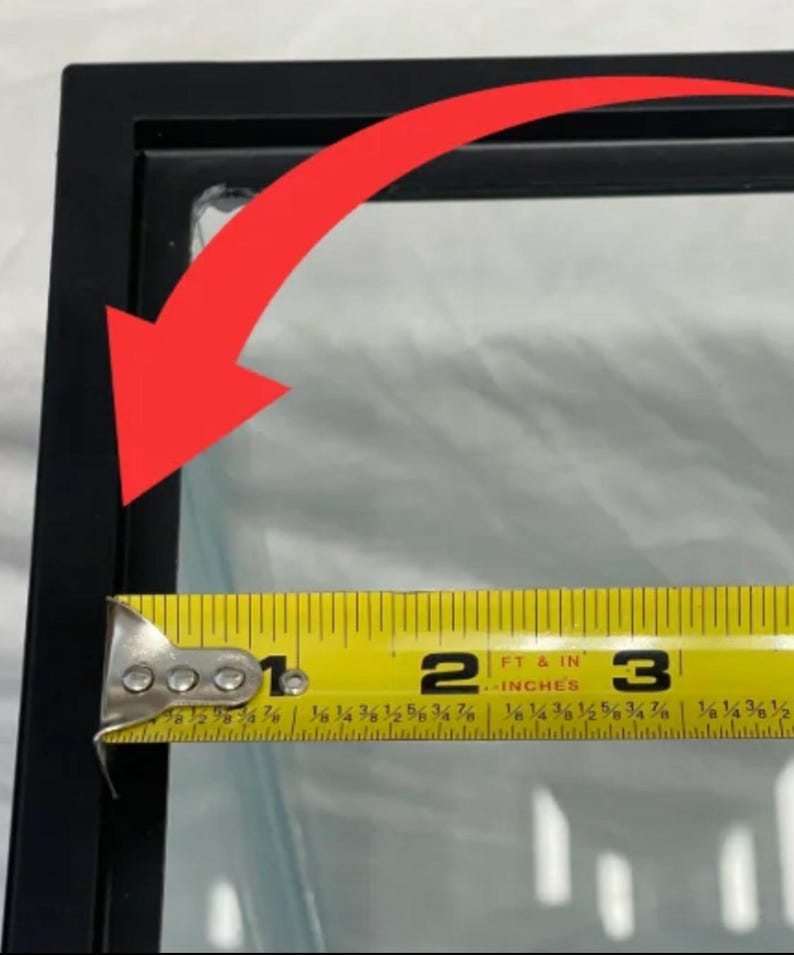May include: A black picture frame with a clear glass pane. A yellow tape measure shows the frame's width is approximately 3 inches. The frame is on a white surface.