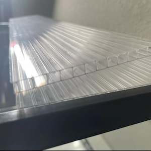 May include: Clear, corrugated plastic sheets stacked on a black surface. The sheets have a ribbed texture, and the light passes through them, creating a translucent effect. The sheets appear to be new and unused.