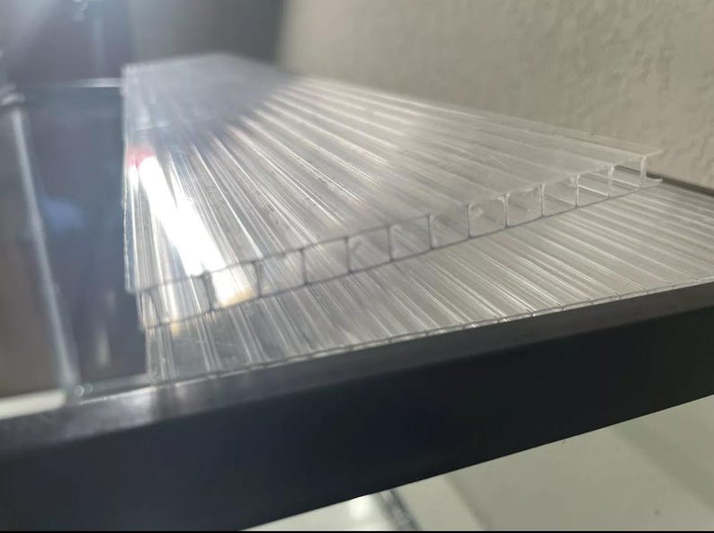 May include: Clear, corrugated plastic sheets stacked on a black surface. The sheets have a ribbed texture, and the light passes through them, creating a translucent effect. The sheets appear to be new and unused.