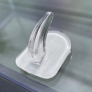 May include: A clear, transparent, plastic object with a curved, fin-like shape rising from a rectangular base. The base has a slightly rounded shape. The object is smooth and reflective, with visible internal details.