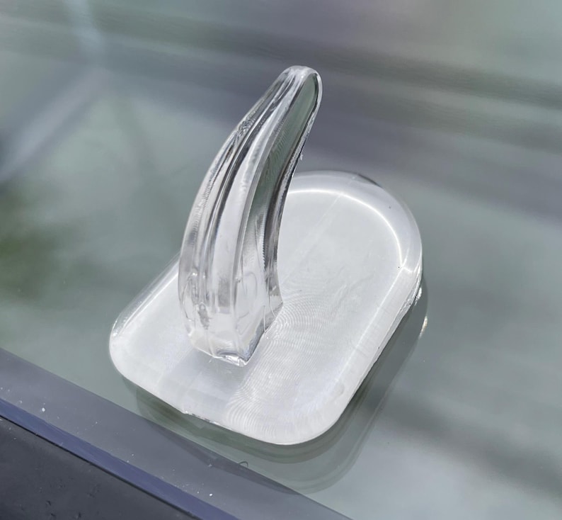 May include: A clear, transparent, plastic object with a curved, fin-like shape rising from a rectangular base. The base has a slightly rounded shape. The object is smooth and reflective, with visible internal details.