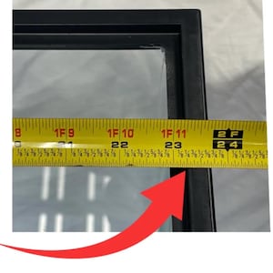 May include: A close-up of a black rectangular frame with a clear glass panel. A yellow measuring tape is placed across the frame, showing a measurement of approximately 24 inches.