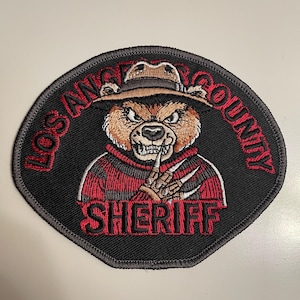 May include: A black and red embroidered patch with a cartoon bear wearing a hat and a red and black striped sweater. The bear has a menacing expression and is holding a clawed hand up to its mouth. The text "Los Angeles County Sheriff" is embroidered around the bear.
