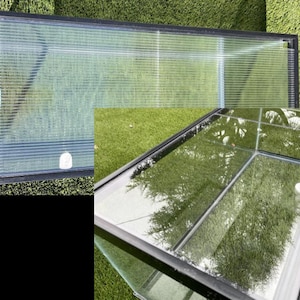 May include: A rectangular glass terrarium with a black frame, set on green grass. The transparent panels allow a view of the grass and reflections. A small white object is visible inside.