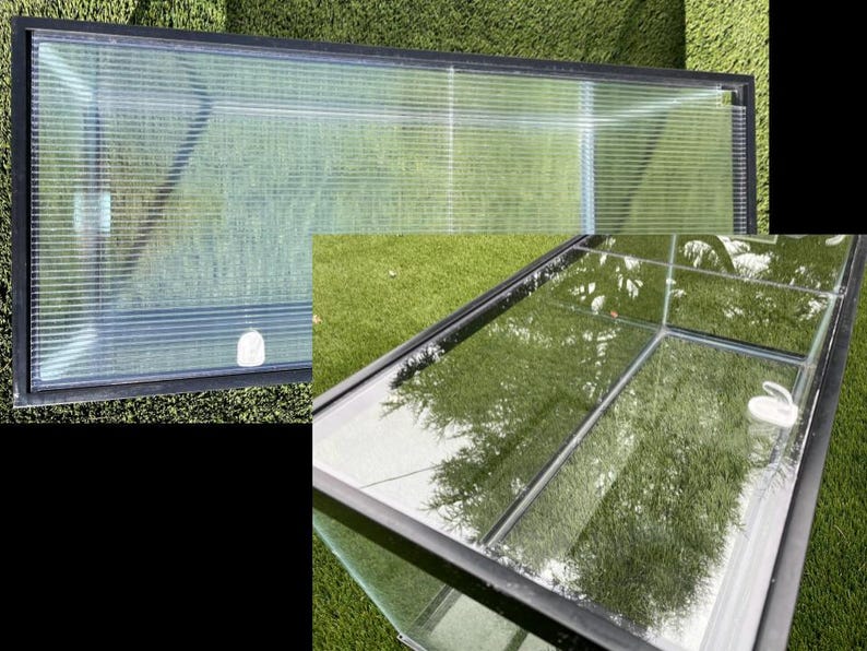 May include: A rectangular glass terrarium with a black frame, set on green grass. The transparent panels allow a view of the grass and reflections. A small white object is visible inside.