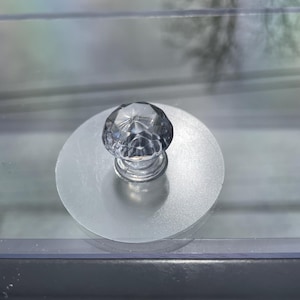 May include: A clear, faceted crystal knob with a silver base is centered on a frosted, circular disc. The knob is likely for a drawer or cabinet, adding a touch of elegance. The image is a close-up shot.