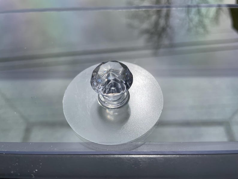 May include: A clear, faceted crystal knob with a silver base is centered on a frosted, circular disc. The knob is likely for a drawer or cabinet, adding a touch of elegance. The image is a close-up shot.