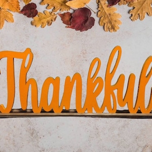 May include: Orange wooden sign with the word "Thankful" in a cursive font. The sign is on a wooden base and is surrounded by autumn leaves.