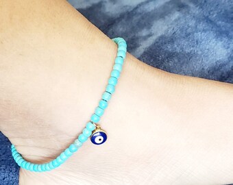 May include: A turquoise beaded anklet with a gold charm featuring an evil eye design. The anklet is worn on an ankle, showcasing the delicate beads and the charm's intricate details. A stylish accessory.