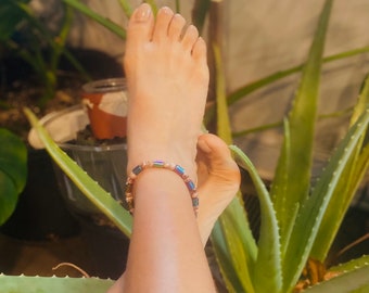 May include: Ankle bracelet with iridescent and tan beads. The bracelet is worn on an ankle, with aloe vera plants in the background. The bracelet has a mix of colours, including blue, pink, and gold.