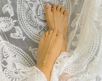 Könnte beinhalten: Nahaufnahme von Füßen mit zartem, goldenem Fußschmuck. Der Schmuck besteht aus einer Kette, die mit einem Zehenring und einem Armband verbunden ist, was einen eleganten, minimalistischen Look ergibt. Die Füße ruhen auf weißem Spitzenstoff mit floralem Muster.