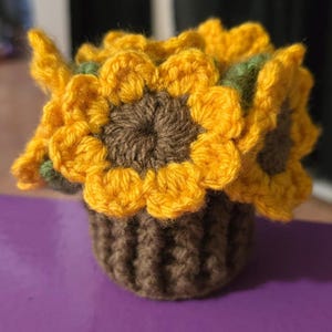 May include: A crocheted sunflower arrangement in a brown pot. The sunflowers have yellow petals, brown centers, and green leaves. The pot is a dark brown color. The arrangement is on a purple surface.