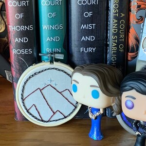 May include: A collection of five fantasy novels with the title "A Court of" on the spines. The books are arranged from left to right: "A Court of Thorns and Roses", "A Court of Wings and Ruin", "A Court of Mist and Fury", "A Court of Frost and Starlight", and a fifth book with an unreadable title.  There are two small figurines in front of the books. One is a blue dress with a crown and the other is a black and grey figure with a purple eye.