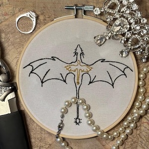 May include: A white embroidery hoop with a black and gold embroidered dragon design. The dragon is facing away from the viewer with its wings spread out. The embroidery is surrounded by a pearl necklace.