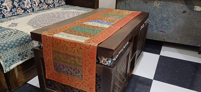 以下が含まれることがあります： カラフルな模様のテーブルランナーが置かれた、長方形の濃い茶色の木製コーヒーテーブル。 ランナーは、オレンジ、青、緑、金などのさまざまな色とパターンでパッチワークデザインになっています。 テーブルの側面には彫刻が施されています。 テーブルランナーは約150cmです。