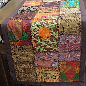 May include: A colourful patchwork table runner featuring embroidered floral designs in shades of brown, green, orange, and purple. The runner is made from various fabrics and textures, creating a unique and eclectic look.
