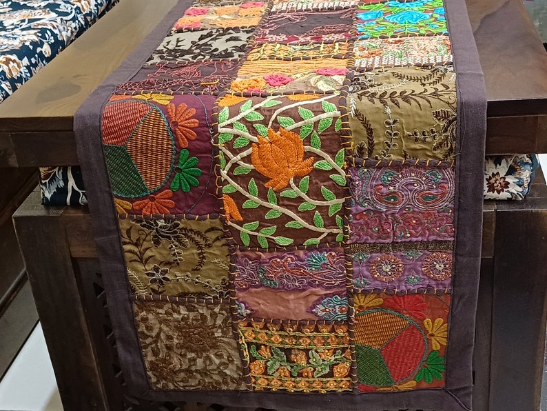 May include: A colourful patchwork table runner featuring embroidered floral designs in shades of brown, green, orange, and purple. The runner is made from various fabrics and textures, creating a unique and eclectic look.