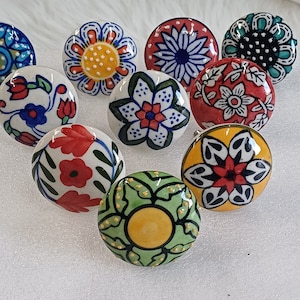 Könnte beinhalten: Zehn Keramik-Knöpfe mit bunten Blumenmustern. Die Knöpfe sind rund und haben einen weißen Hintergrund. Die Muster umfassen Rot, Blau, Grün, Gelb und Schwarz.