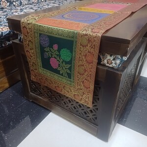 May include: A dark wood coffee table with a decorative embroidered table runner featuring floral designs in shades of green, pink, and blue. The table runner has a gold border and is placed on top of a dark wood surface.