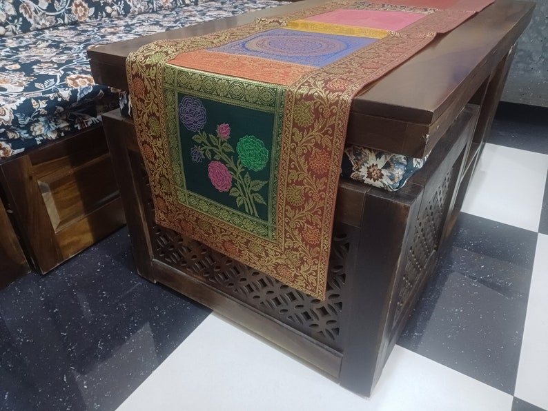May include: A dark wood coffee table with a decorative embroidered table runner featuring floral designs in shades of green, pink, and blue. The table runner has a gold border and is placed on top of a dark wood surface.