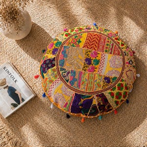 May include: A round, patchwork floor cushion with a vibrant mix of colors, including yellow, pink, and purple. The cushion features a central circular design and is adorned with small, colorful tassels around the edges. A magazine with the word "KINFOLK" is visible.