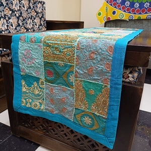 K&ouml;nnte beinhalten: Ein t&uuml;rkisfarbener Tischl&auml;ufer mit Patchwork-Muster in verschiedenen Blaut&ouml;nen, Gr&uuml;n- und Goldt&ouml;nen. Der L&auml;ufer ist aus Stoff mit Stickereien und hat einen Fransenrand.