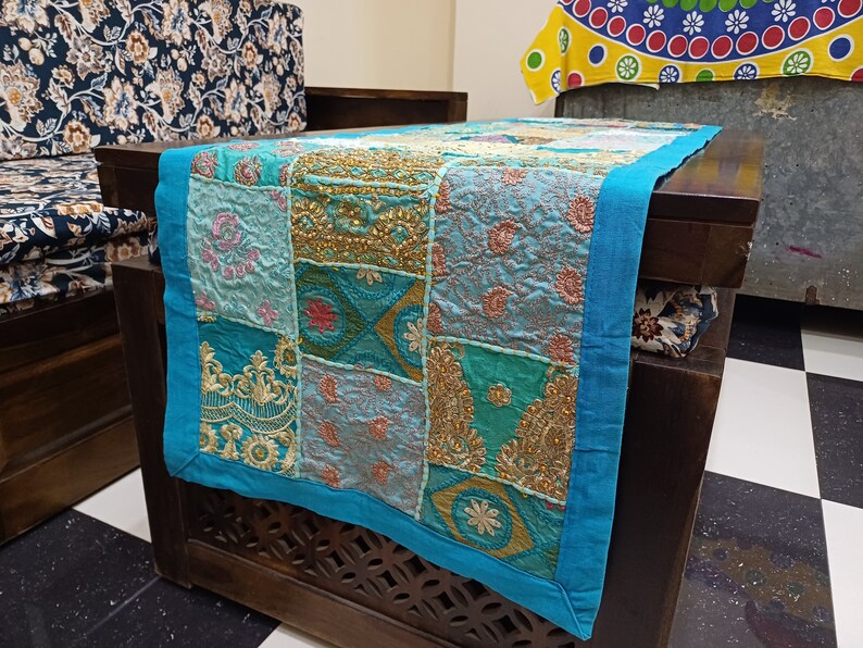 K&ouml;nnte beinhalten: Ein t&uuml;rkisfarbener Tischl&auml;ufer mit Patchwork-Muster in verschiedenen Blaut&ouml;nen, Gr&uuml;n- und Goldt&ouml;nen. Der L&auml;ufer ist aus Stoff mit Stickereien und hat einen Fransenrand.