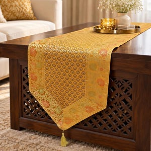 May include: A rectangular gold table runner featuring a repeating floral pattern in shades of orange, green, and yellow. The fabric has a woven texture with a pointed end finished with a single gold tassel hanging from the center point.