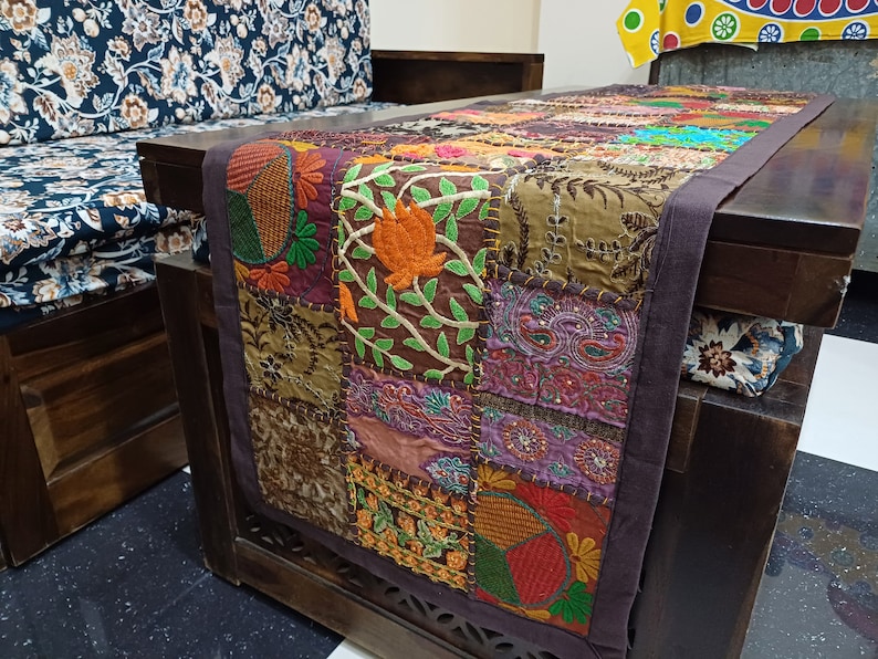 May include: A brown table runner with a patchwork design featuring colourful embroidered floral patterns. The runner is made of fabric and has a dark brown border.
