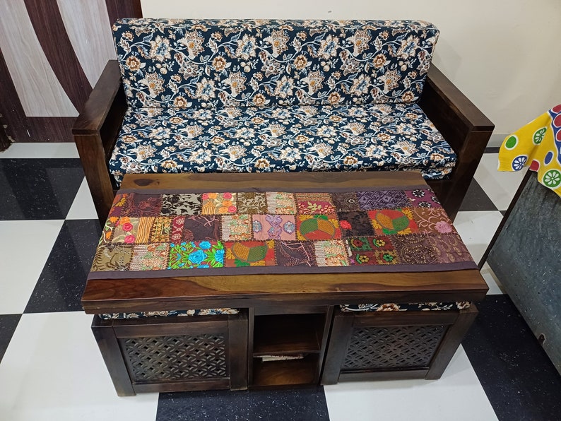 May include: A dark wood sofa with a floral patterned fabric cushion and a matching dark wood coffee table with a colourful patchwork runner. The coffee table has two storage compartments with carved wooden doors and a shelf in the middle.